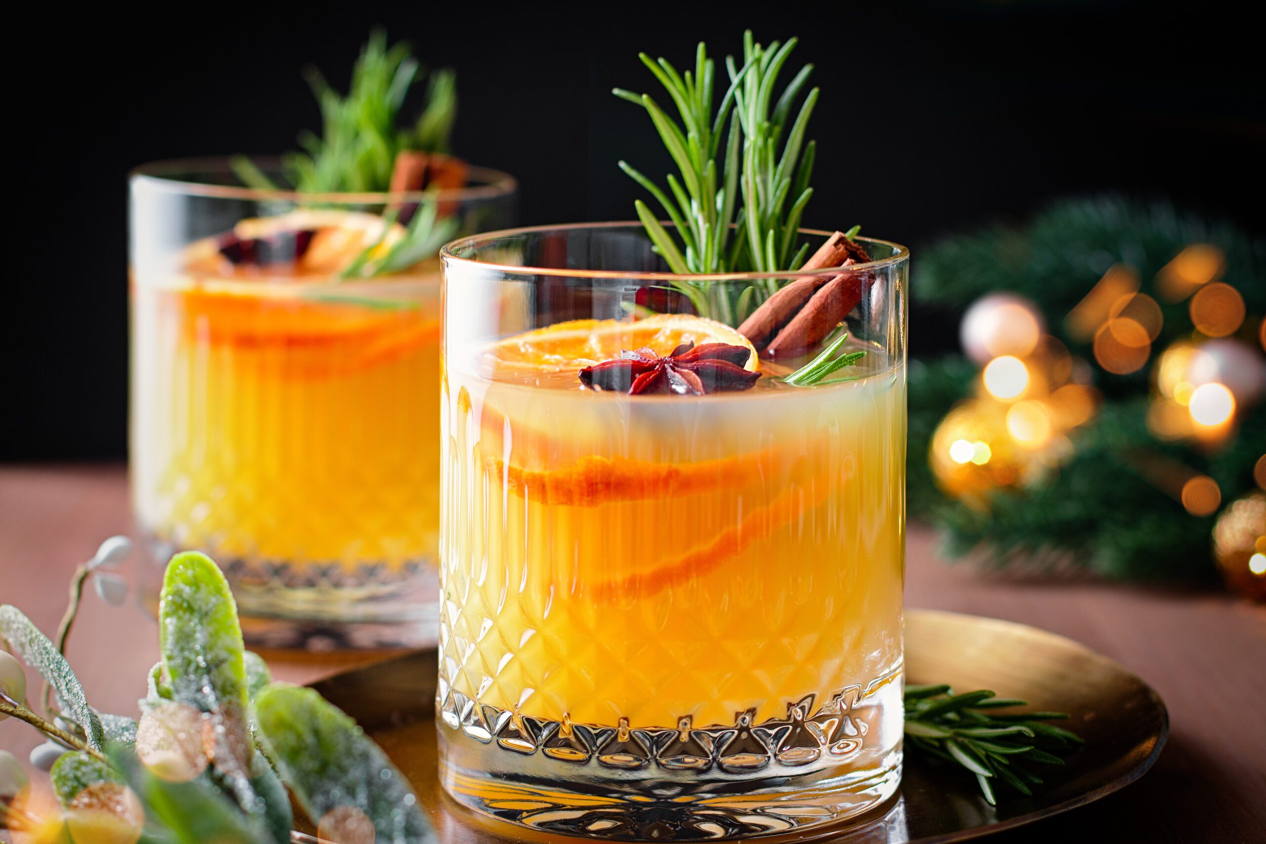 Christmas or winter warming bourbon old fashioned with oranges, cinnamon and rosemary on wooden table with tree branches and golden decor, top view
