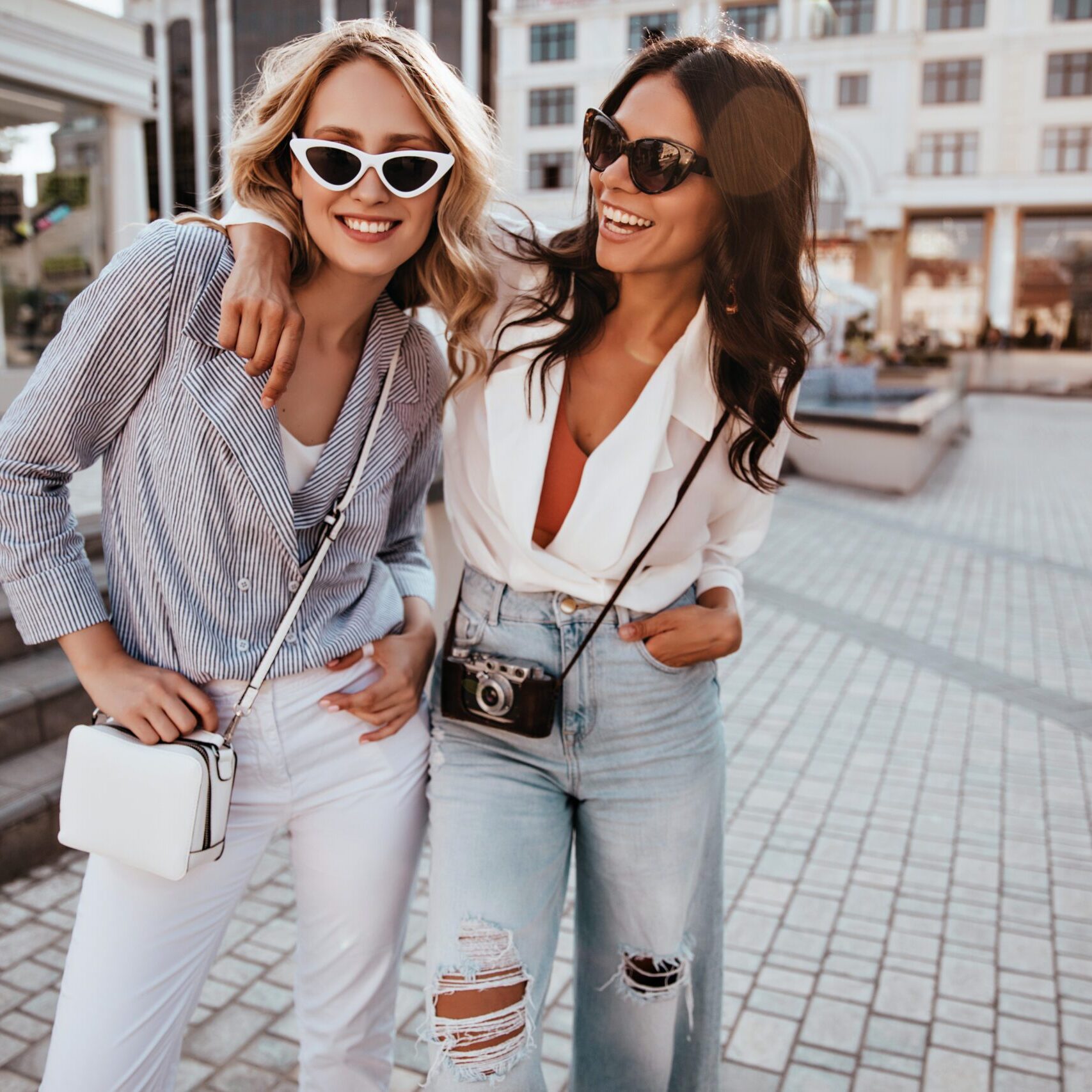 Confident blonde girl in sunglasses enjoying outdoor photoshoot with best friend. Stunning dark-haired female model embracing sister on the street.