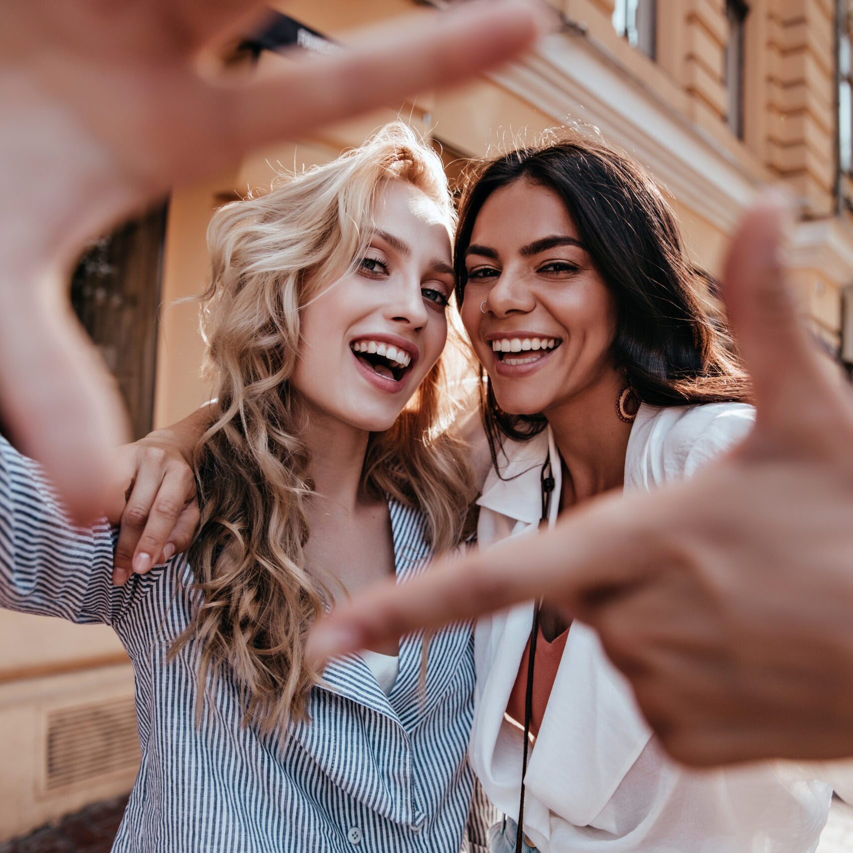 Laughing blithesome woman with dark hair walking down the street with friend. Carefree blonde girl posing on city background.